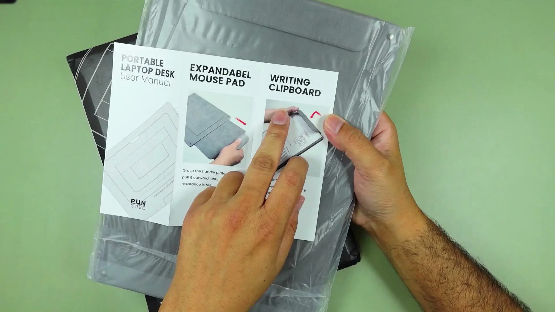 Hands consulting PUNCUBE's printed user manual for its portable laptop desk, showing the mousepad and clipboard fold-out functions