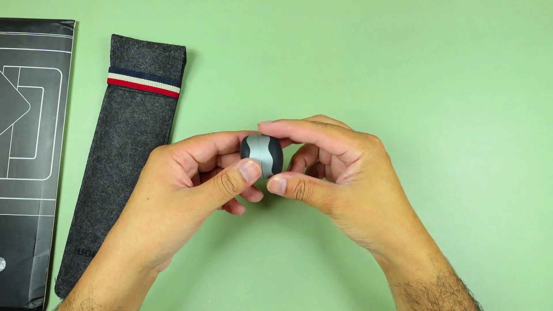Two hands holding the Hagibis magnetic laptop stand folded down into its compact pebble form, on a green test mat