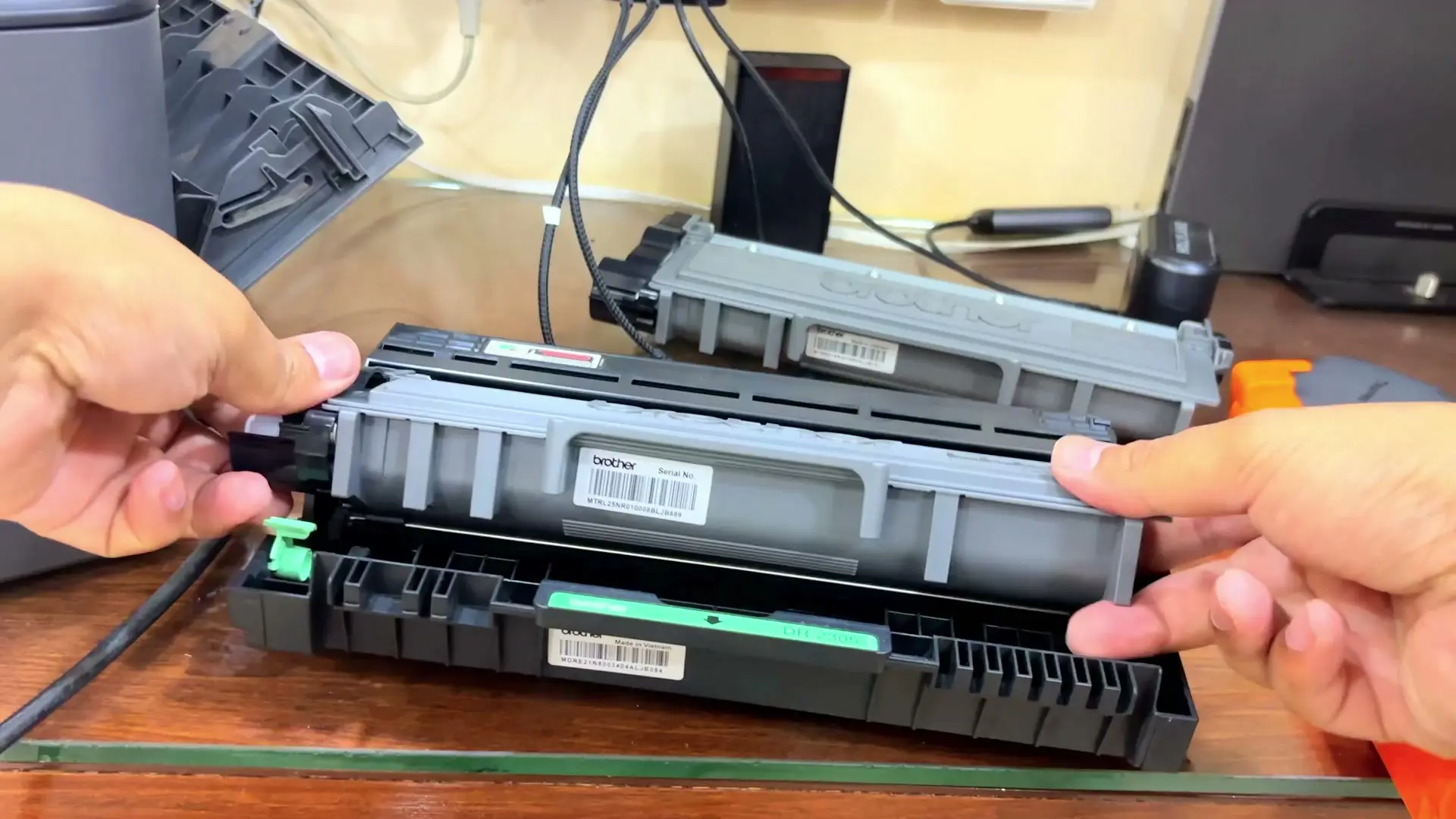 Two hands holding a Brother toner cartridge, Brother logo visible, above a drum unit next to the open printer