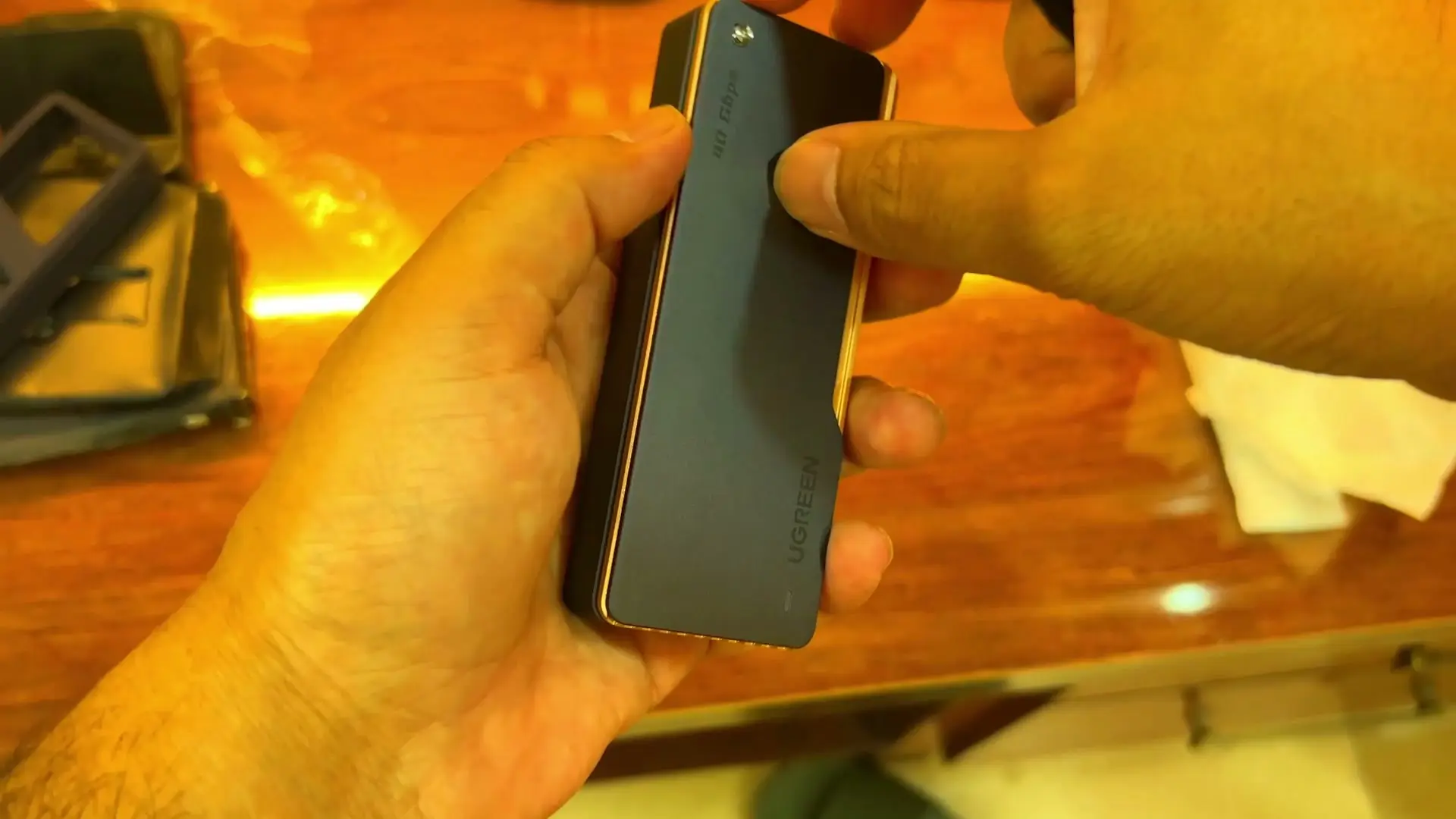 A hand holding the UGREEN 40Gbps M.2 NVMe enclosure, its dark metal body showing the UGREEN name and a 40 Gbps mark, on a wooden desk