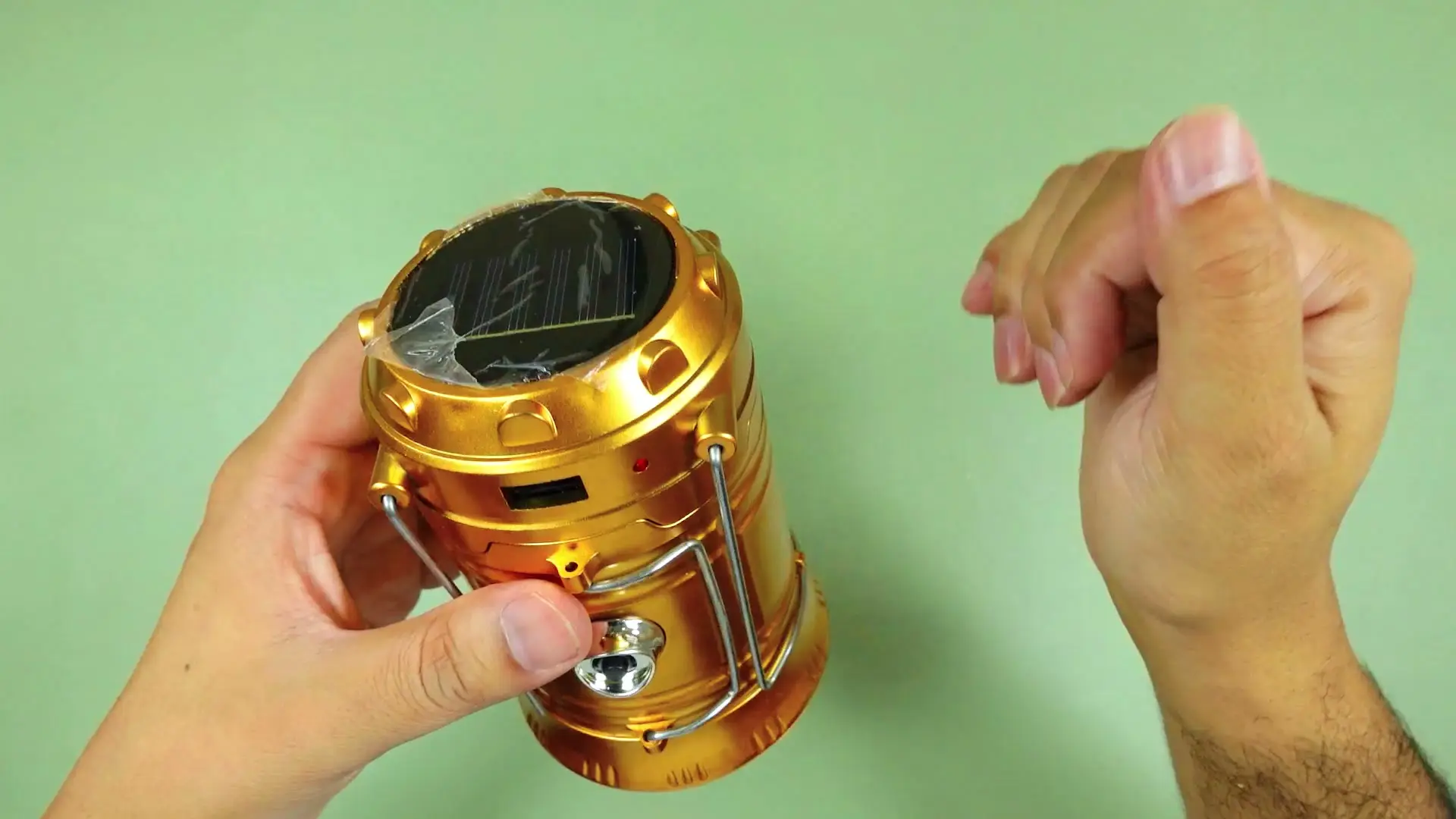 Hands holding the freshly unboxed gold collapsible solar camping lantern, solar panel still covered in factory protective film