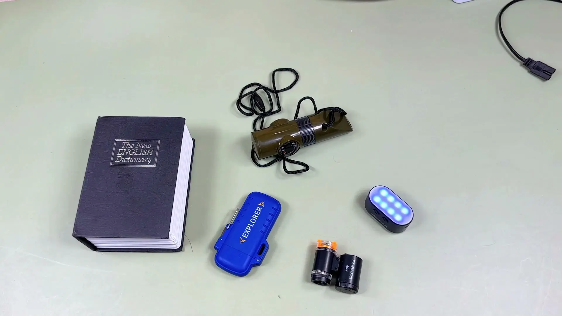 All five tested gadgets laid out together on the test surface: the dictionary book safe, USB arc lighter, pocket microscope, survival whistle, and AOCHUAN magnetic fill light