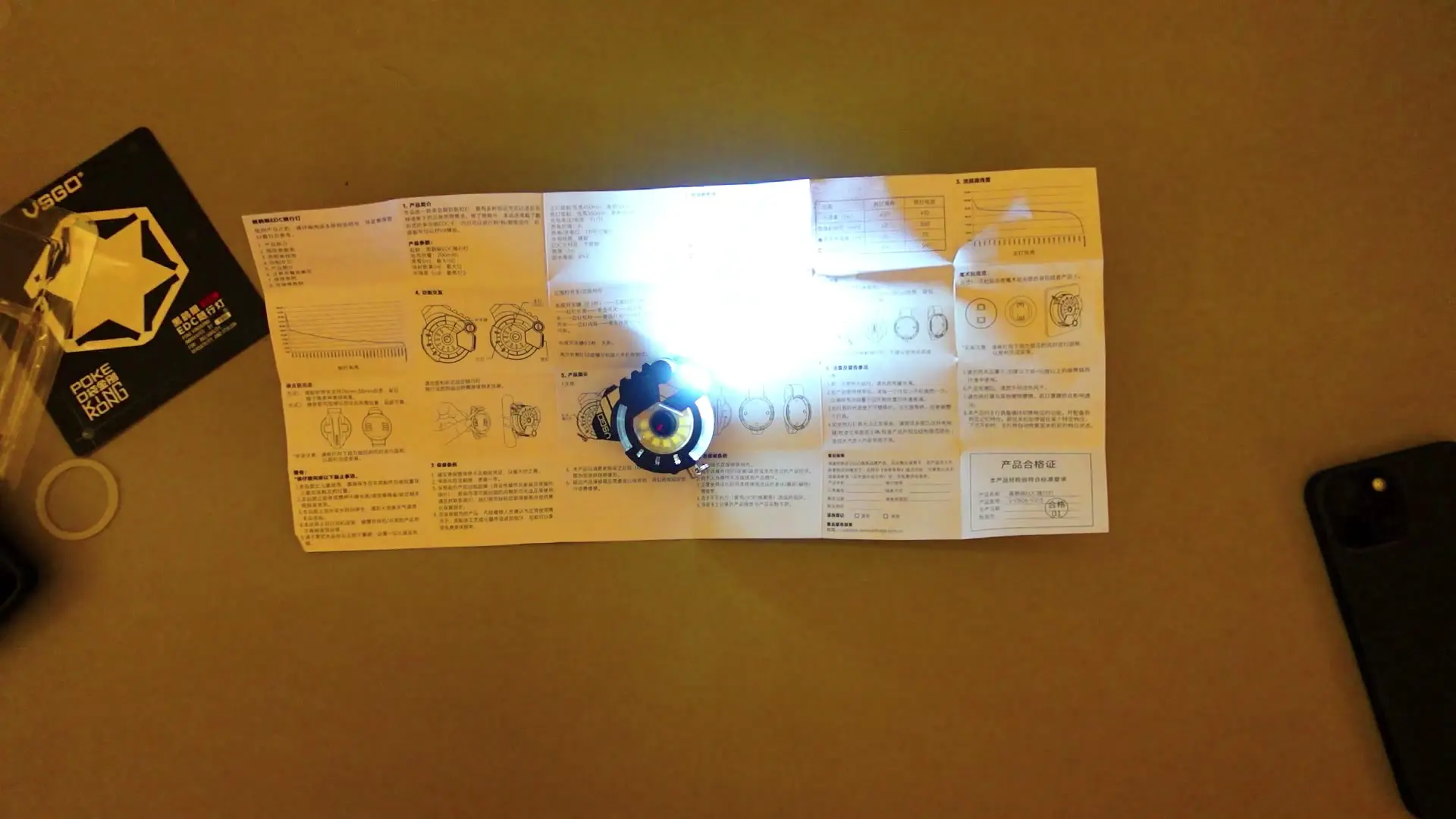 Tiny black VSGO EDC flashlight turned on with a bright white beam shining across an instruction manual